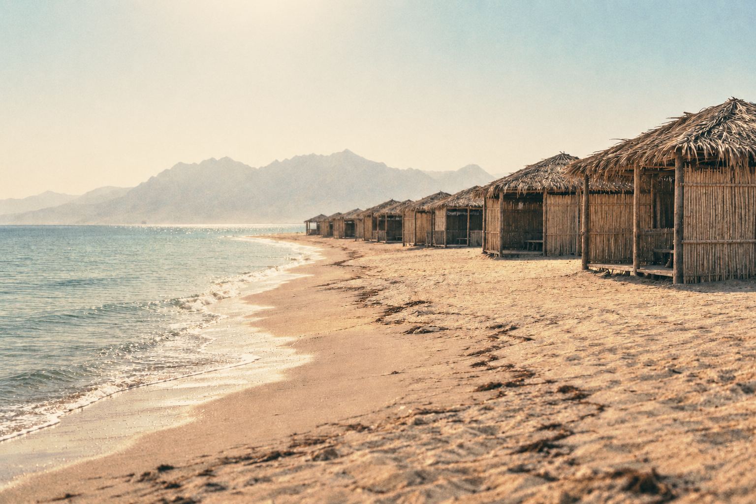 Thatched palm-leaf beach huts in a row on the sand under a hazy noon sun, the sea calm and pale.