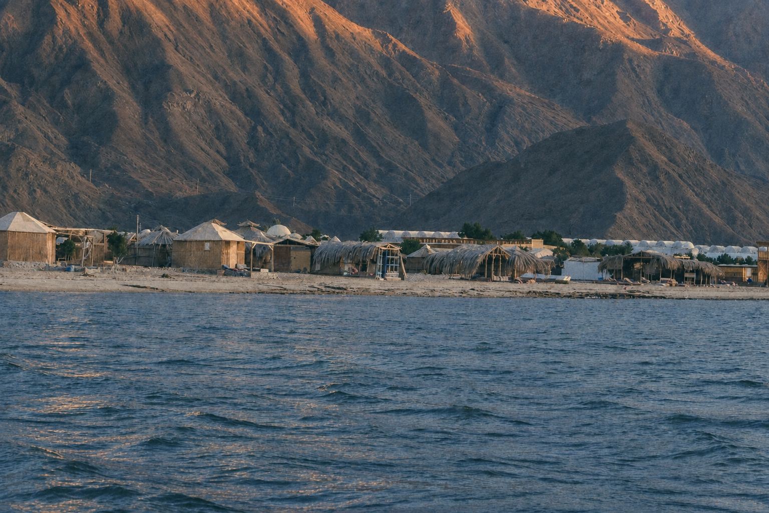 Low sandy-roofed camp huts in late-afternoon shade at the foot of terracotta-red granite mountains, beside a calm sea.