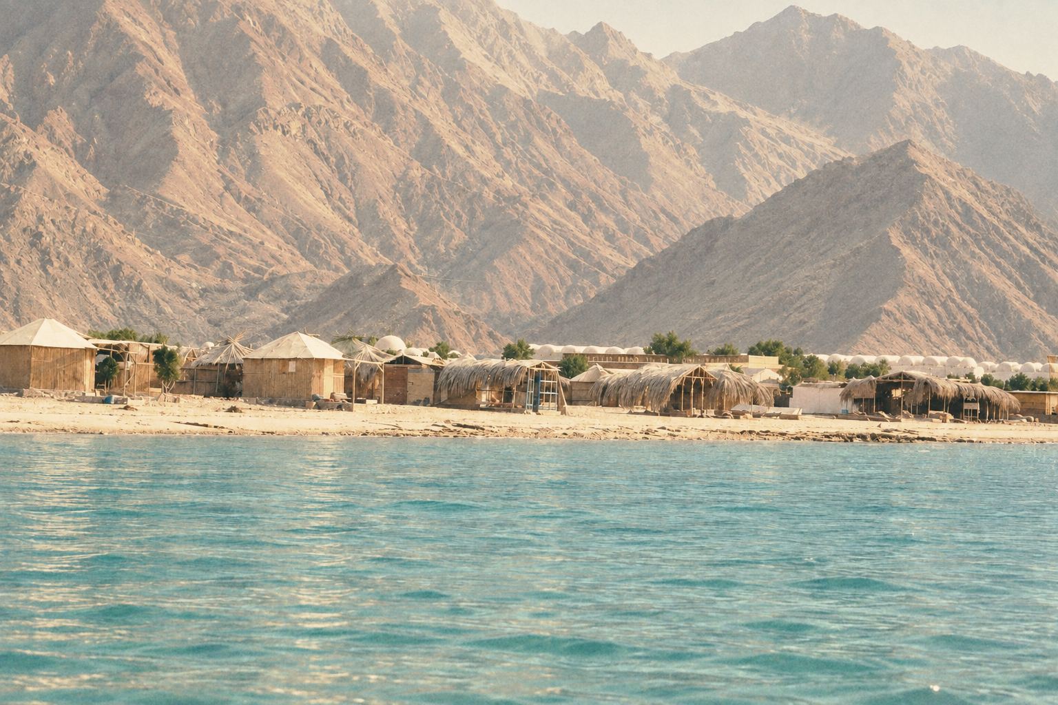 Low huts beneath a huge granite mountain beside a pale sea in bright midday haze.