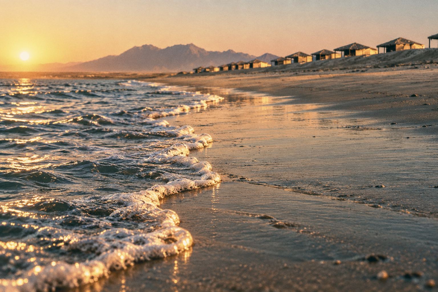 Foam lace from a wave runs across wet sand at golden hour, with distant huts and hazy mountains.